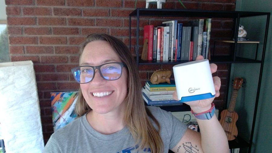 Smiling and holding up a white and blue AirGradient air quality monitor in front of a brick wall and bookshelf