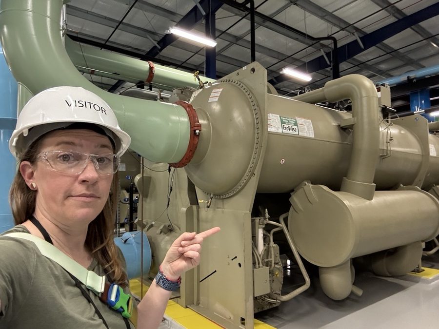 Pointing at a large water-cooled chiller while wearing a white visitor hard hat and safety glasses