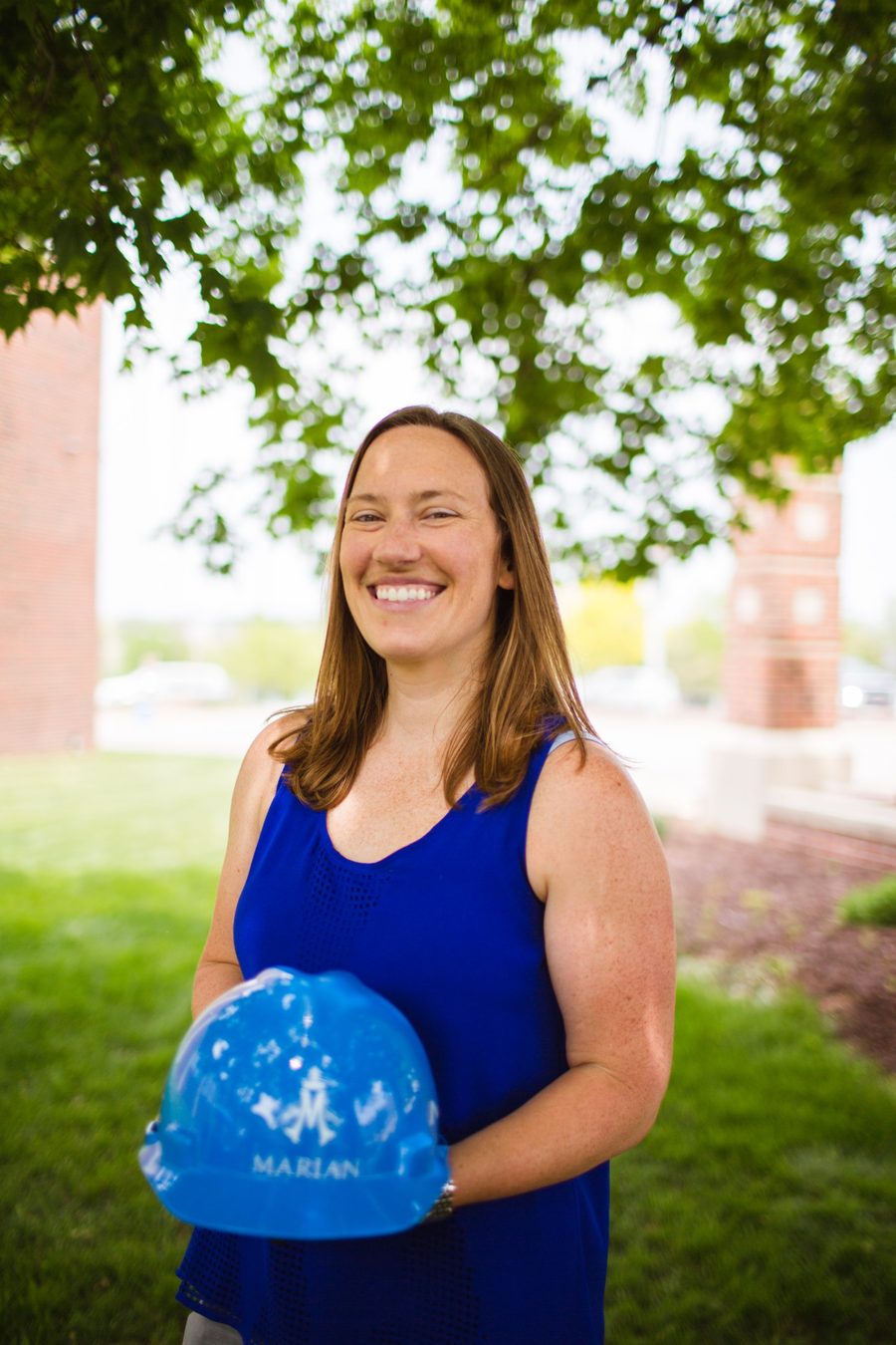 Smiling outdoors under a tree, holding a blue Marian High School hard hat